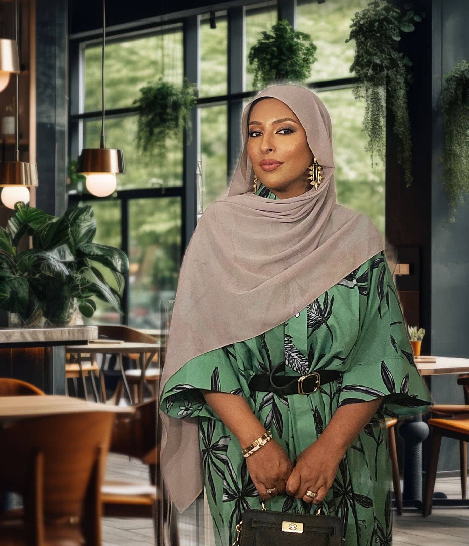 Woman in a green patterned dress and beige hijab standing in a modern cafe.